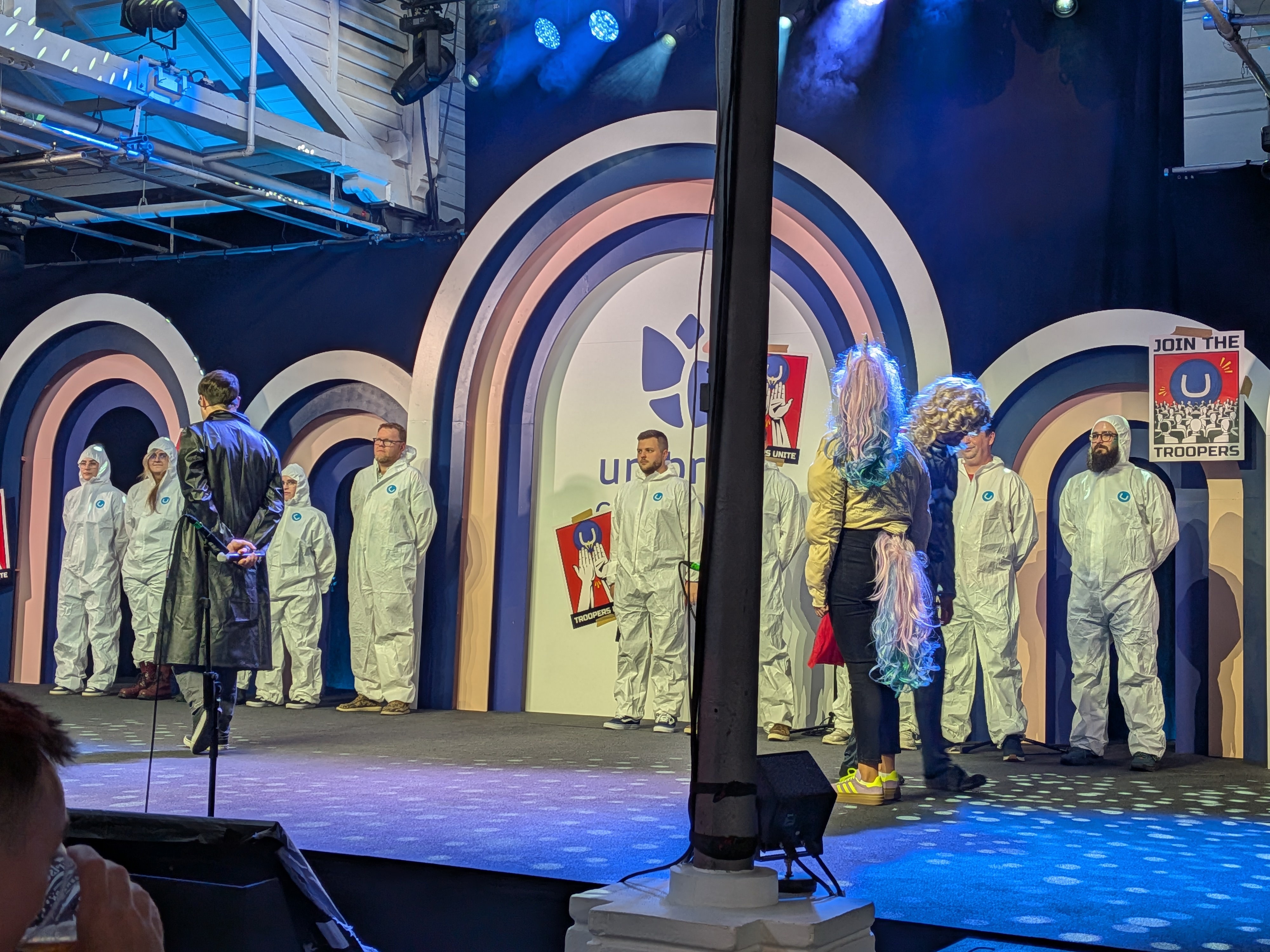 A group of performers in white coveralls stand across a stage with colorful arch backdrops while two costumed presenters face them under blue stage lights.