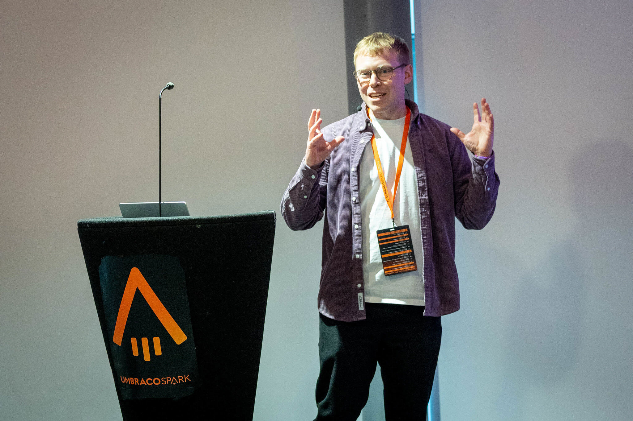 A speaker in a purple shirt and orange lanyard gestures as he talks from beside the Umbraco Spark lectern at Spark 2026.